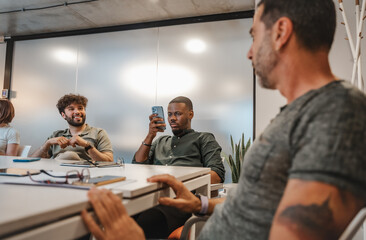 Fototapeta premium Good looking black male person scrolling on his phone while his colleagues are having a conversation