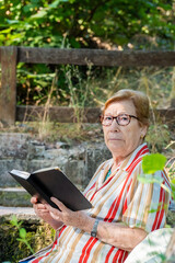  An older woman looking at the camera with a book in her hands, rests relaxed in a park