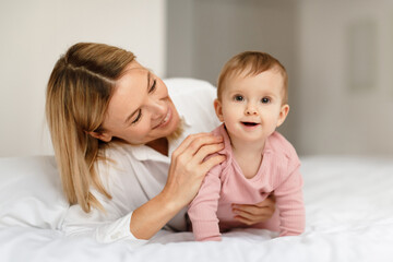 Loving mother talking and playing to her infant child girl, kid crawling and smiling at camera on bed in bedroom