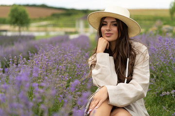 a beautiful girl with long hair and wearing a hat poses outdoors in a field of lavender.