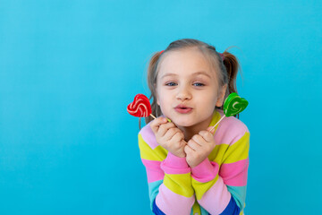 portrait of a joyful little girl with lollipops in a striped jacket, a large candy on a stick. The concept of sweets and confectionery. Blue background, photo studio, place for text