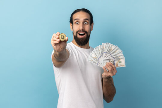 Portrait of surprised man with beard wearing white T-shirt showing bitcoin and big fan of dollars banknotes, e-commerce, crypto currency. Indoor studio shot isolated on blue background.