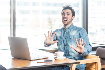 Fototapeta na wymiar Portrait of shocked panicked scared young man freelancer in blue jeans shirt working on laptop, showing stop gesture, looks frighten, screaming. Indoor shot near big window, cafe background.