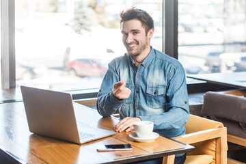 Fototapeta na wymiar Portrait of delighted satisfied smiling bearded young man freelancer in blue jeans shirt working on laptop, pointing finger to camera. Indoor shot near big window, cafe background.