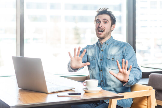 Portrait of shocked panicked scared young man freelancer in blue jeans shirt working on laptop, showing stop gesture, looks frighten, screaming. Indoor shot near big window, cafe background.