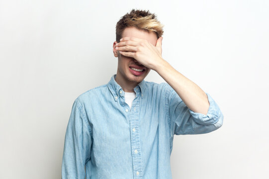 Portrait of shocked scared man wearing denim shirt standing covering his eyes with palm, doesn't want to see something frighten. Indoor studio shot isolated on gray background.