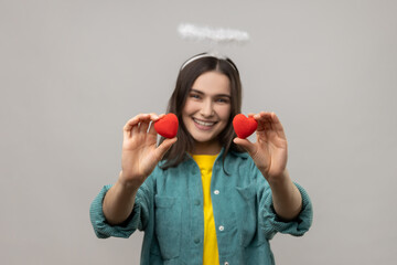 Satisfied young angelic woman with nimb over head, showing to camera two little red hearts, expressing romantic feelings, wearing casual style jacket. Indoor studio shot isolated on gray background.