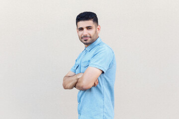 Side view portrait of attractive confident positive man wearing denim shirt holding arms folded, looking at camera, looks happy. Indoor studio shot isolated on gray background.