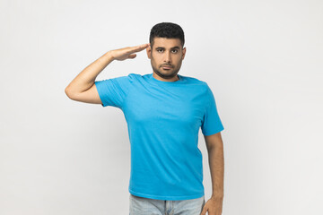 Portrait of serious strict handsome unshaven man wearing blue T- shirt standing with hand near,...