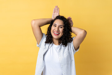 Portrait of happy woman with dark wavy hair making funny bunny ears with hands on head, childish behavior, playful optimistic mood. Indoor studio shot isolated on yellow background.