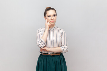 Portrait of pensive thoughtful woman wearing striped shirt and green skirt holding her chin,...