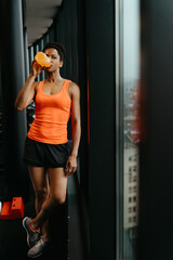 Sports woman in the gym standing next to the window and drinking water from water bottle hydrating her body after workout. Post workout concept.