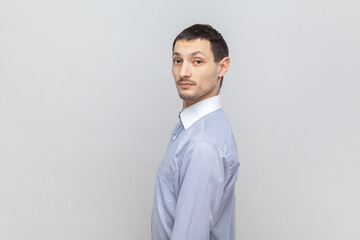 Side view portrait of serious attractive man standing looking at camera with strict concentrated facial expression, wearing light blue shirt. Indoor studio shot isolated on gray background.