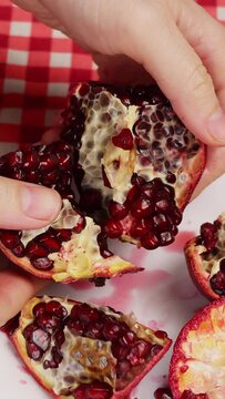 Vertical video,female hands breaking half of ripe pomegranate over white plate to prepare fresh salad with juicy red pomegranate seeds.