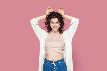 Portrait of funny childish woman with curly hair wearing casual style outfit having fun, making bunny ears with palms, looking smiling at camera. Indoor studio shot isolated on pink background.