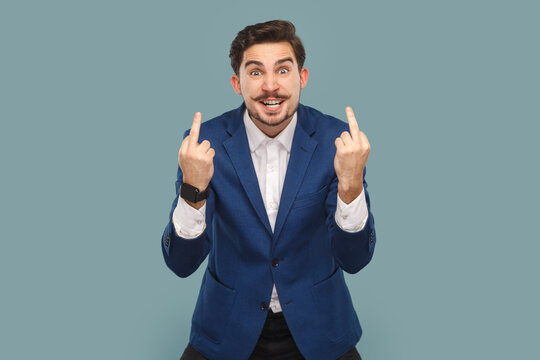 Rude impolite handsome man with mustache standing showing middle fingers, looking at camera with clenched teeth, wearing white shirt and jacket. Indoor studio shot isolated on light blue background.