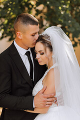 A stylish groom in a black suit and a cute bride in a white dress with a long veil are hugging in a park. Wedding portrait of smiling and happy newlyweds.