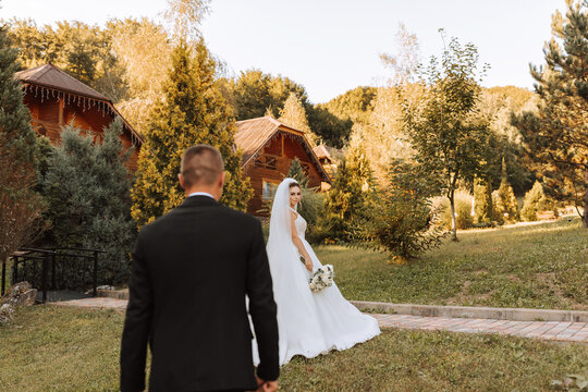 A stylish groom in a black suit and a cute bride in a white dress with a long veil are hugging and walking near green tall trees. Wedding portrait of smiling and happy newlyweds.