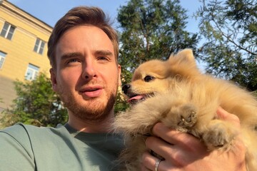 Young man holding on hands his Pomeranian Spitz dog, walking with cute puppy at summer day 