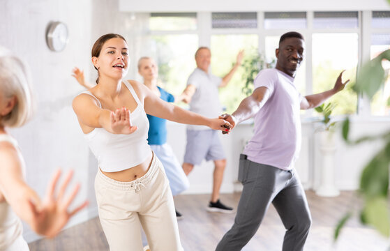 In fitness studio, young girl teacher paired with african man dancing Latin American dance. Clients of different ages are engaged in fitness in background