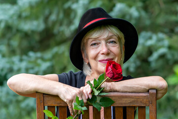 A happy aged woman, wearing a black hat, holding a gifted red rose against the background of nature.