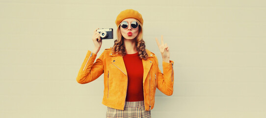 Fashionable autumn color style outfit, stylish young woman photographer with film camera, model posing wearing orange french beret hat, jacket, sunglasses on gray background