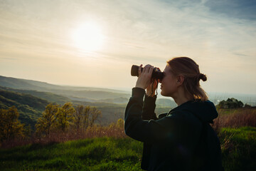 A hiker girl looks through binoculars at nature and birds standing on the top of a mountain in the bright rays of setting sun against the background of beautiful spring nature and cloudy blue sky - Powered by Adobe