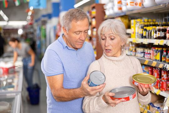 Interested Senior Woman And Man Choosing Together Canned Food Products On Shelves In Supermarket, Examining Labels On Tin Cans And Discussing Before Making Selection..