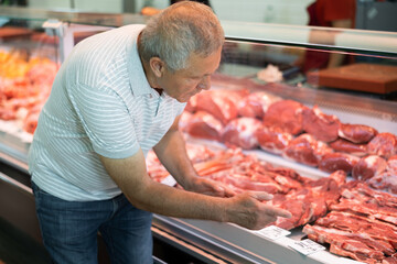 Elderly man buyer of meat department of grocery store, looking at meat in refrigerator window. Male...