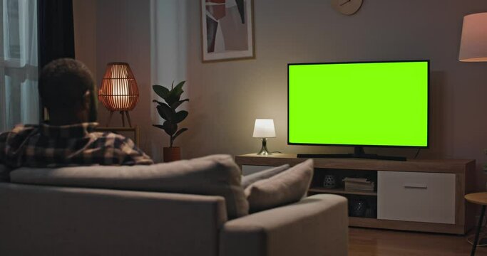 Man sitting at sofa in modern appartment. Back view of male sitting in front of TV. African American man relaxing sitting at couch at well designed room. Male looking at green chroma key screen of TV.