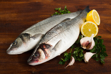 Two whole fresh European basses lying on wooden surface with lemon, parsley and garlic..