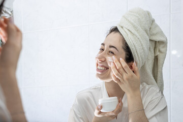 Beautiful woman applies cream on her face after a relaxing bath. Concept of personal care, and...