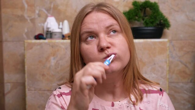 Woman with long hair in pajamas brushes her teeth in toilet with curiosity inspects bathroom, silly expression on her face hums haunting melody. Lady with a toothbrush in her hands relaxes to the beat