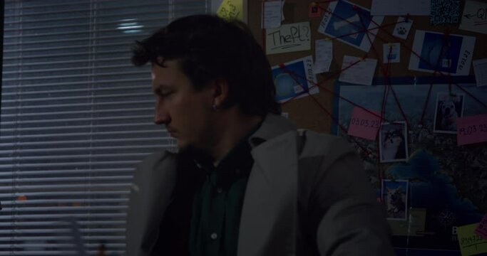 Man criminal investigator sits in office studying evidence on investigation, on wall there is crime board, information on murder, scheme, interconnection Detective workaholic examines new case