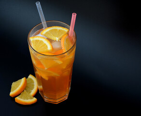 A tall glass of orange juice with ice and straws on a black background, next to pieces of ripe...