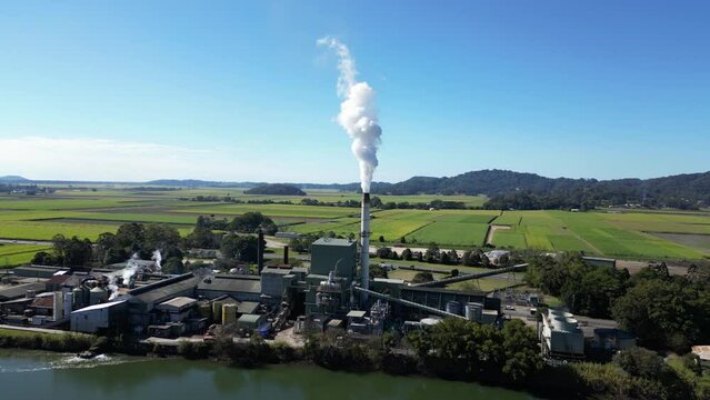 Smoke billows from a sugar mill refinery on the banks of a country river with a mountain backdrop view. Drone