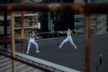 Two fencers woman practice on a rooftop with the city in the background.