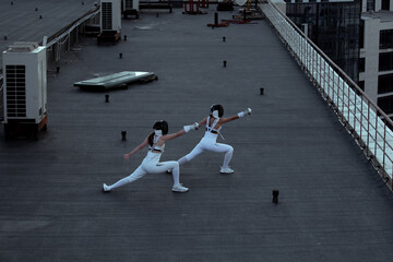 Two fencers woman practice on a rooftop with the city in the background.