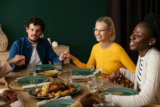 Happy friends holding hands and praying at dining table