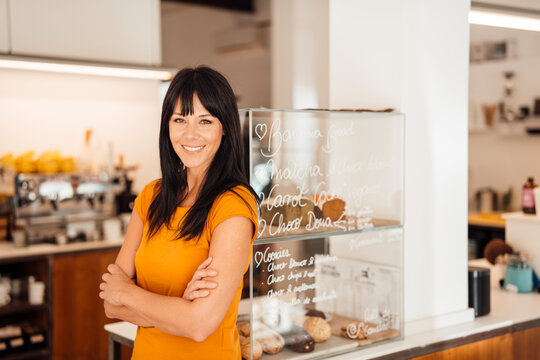 Happy mature woman with arms crossed standing by retail display in cafe