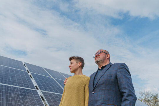 Thoughtful father and son standing next to solar panel under sky