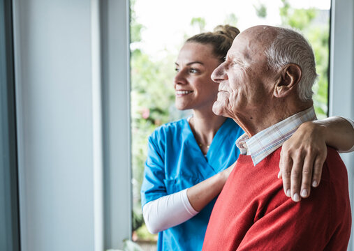 Happy nurse with senior man in front of window at home