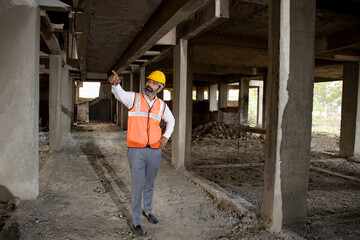 Young Indian male civil engineer or architect wearing helmet and vest holding pointing at construction site.