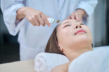 아시아 한국의 젊은 한국여성 모델은 스파 또는 피부관리전문점 또는 피부과 병원에서 침대에 누워서 전문관리사에게 초음파 스크래퍼로 피부관리를 받음.