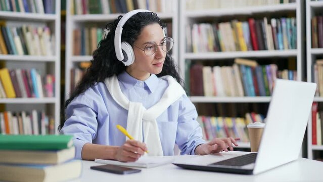 Female student in headphones listens to online e-learning, video course in campus library space. A girl at a distance learning seminar at the university writes in a notebook using a laptop computer