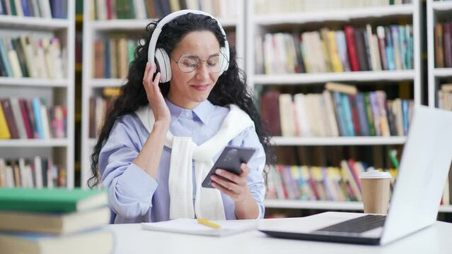 Young smiling female student wearing headphones listening and enjoying music using smartphone sitting in campus library space. Happy satisfied brunette girl turns on a playlist on phone and dances