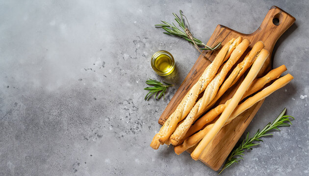 Fresh baked grissini bread sticks on wooden cutting board with olive oil and herbs rosemary and basil over grey concrete textured surface. Top view, copy space.