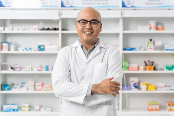 Naklejka na ściany i meble Happy handsome asian male pharmacist wearing eyeglasses and lab coat standing with arms crossed and looking at camera, He feels good, trustworthy and proud of his work in the pharmacy drugstore.