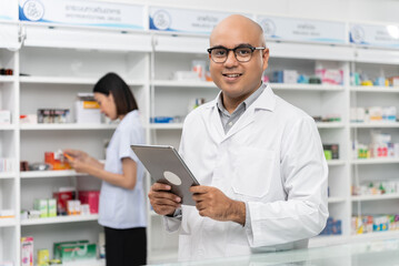 Happy handsome asian male pharmacist wearing eyeglasses and lab coat standing with tablet and looking at camera, He feels good, trustworthy and proud of his work in the pharmacy drugstore.