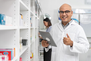 Naklejka na ściany i meble Happy handsome asian male pharmacist wearing eyeglasses and lab coat standing with tablet and thumbs up, He looking at camera feel good, trustworthy and proud of his work in the pharmacy drugstore.